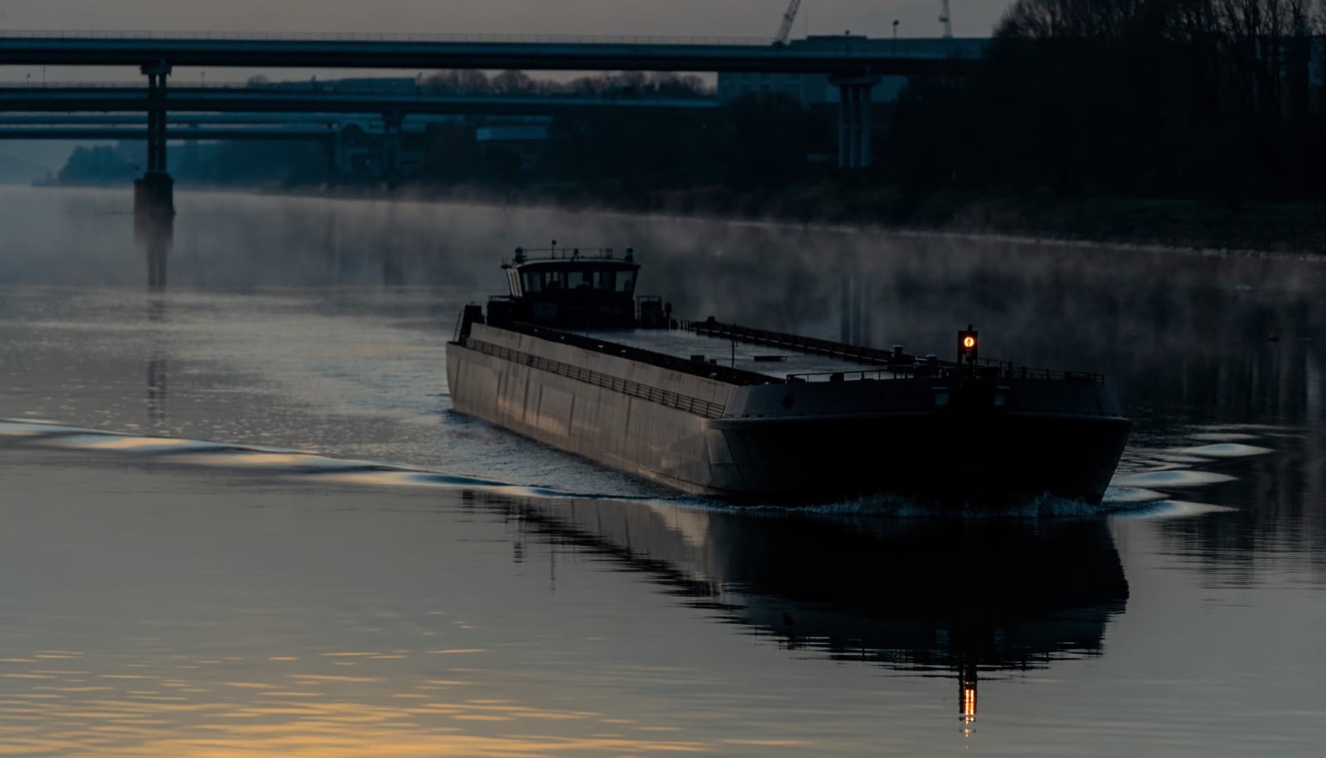 An inland tanker barge under way on a wide river at dawn with mist on the water