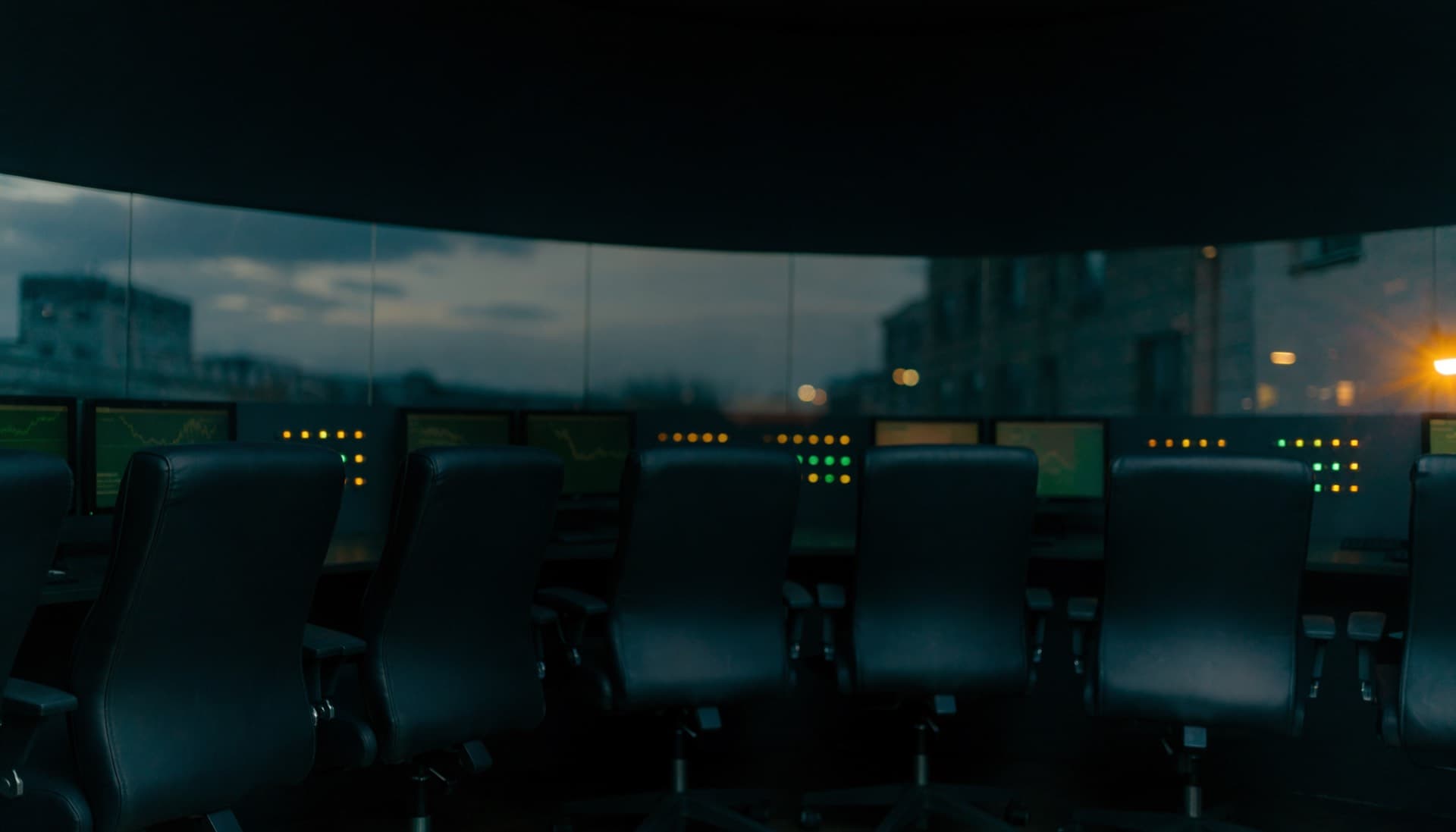 A darkened terminal operations room at night, empty chairs before a bank of dimly glowing monitors
