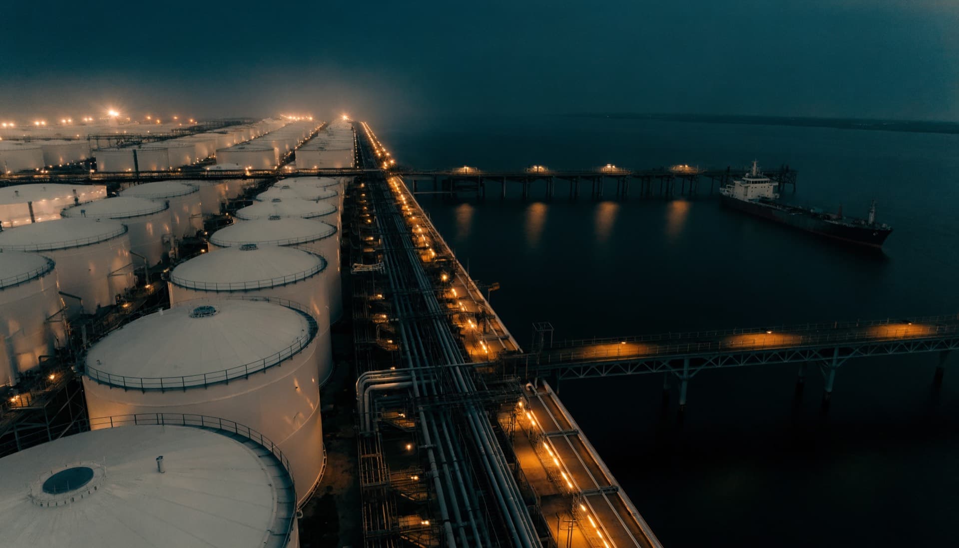 A refined products tank farm and marine loading terminal at blue hour, jetty arms reaching over dark water